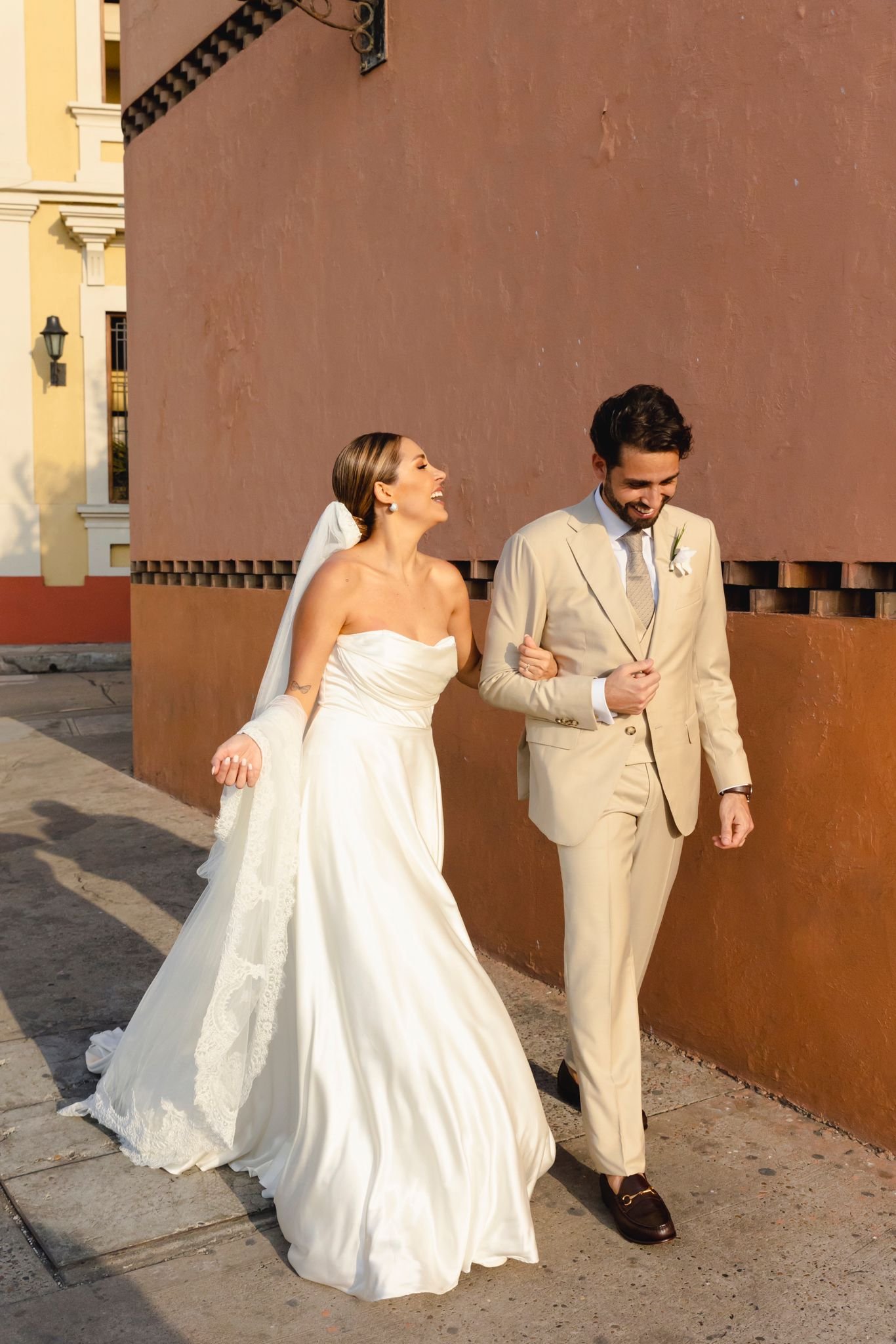 Los novios caminando por una calle colonial con la falda capa de satén en movimiento