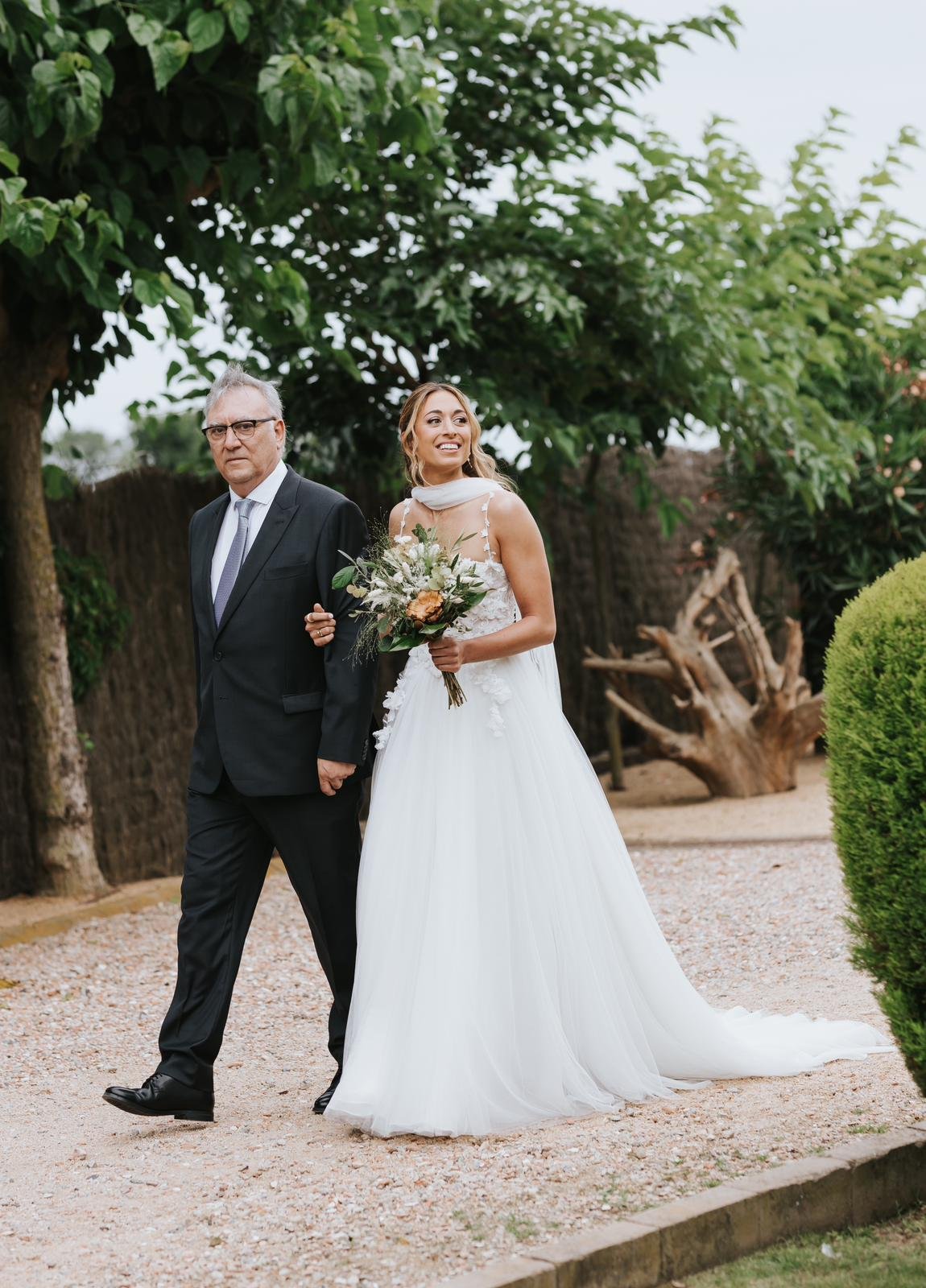 Claudia entrando del brazo de su padre por los jardines de Mas Palau Blanes con su ramo y el vestido de corset floral y falda de tul