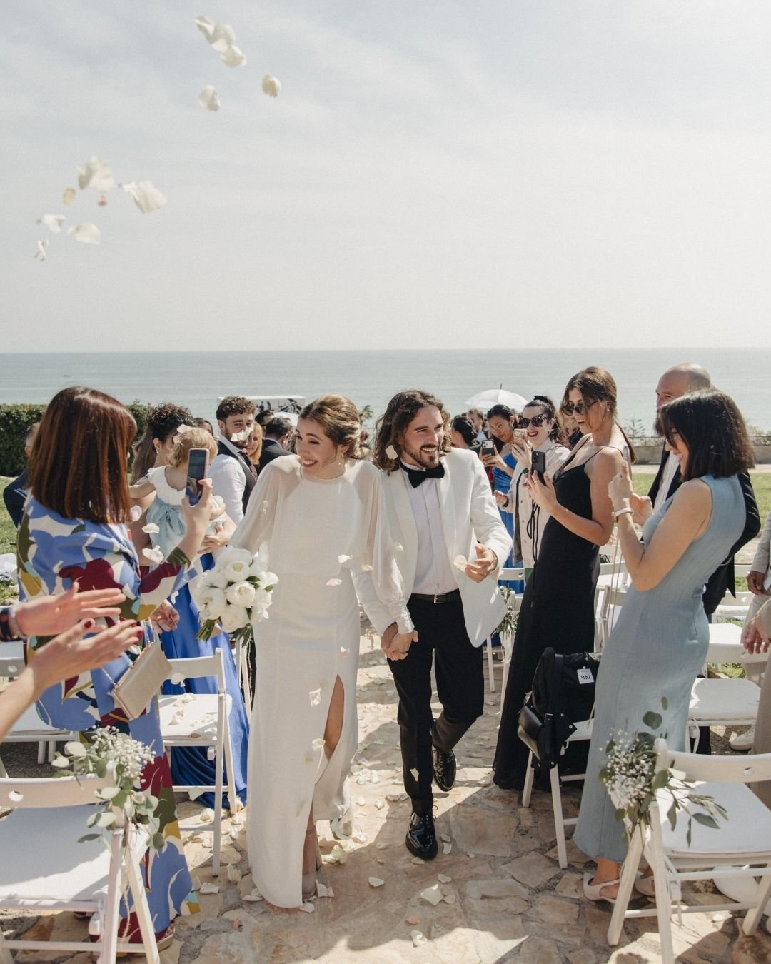 Salida de la ceremonia entre pétalos frente al mar en Santa Romana, vestido de crepé y chaqueta de gasa