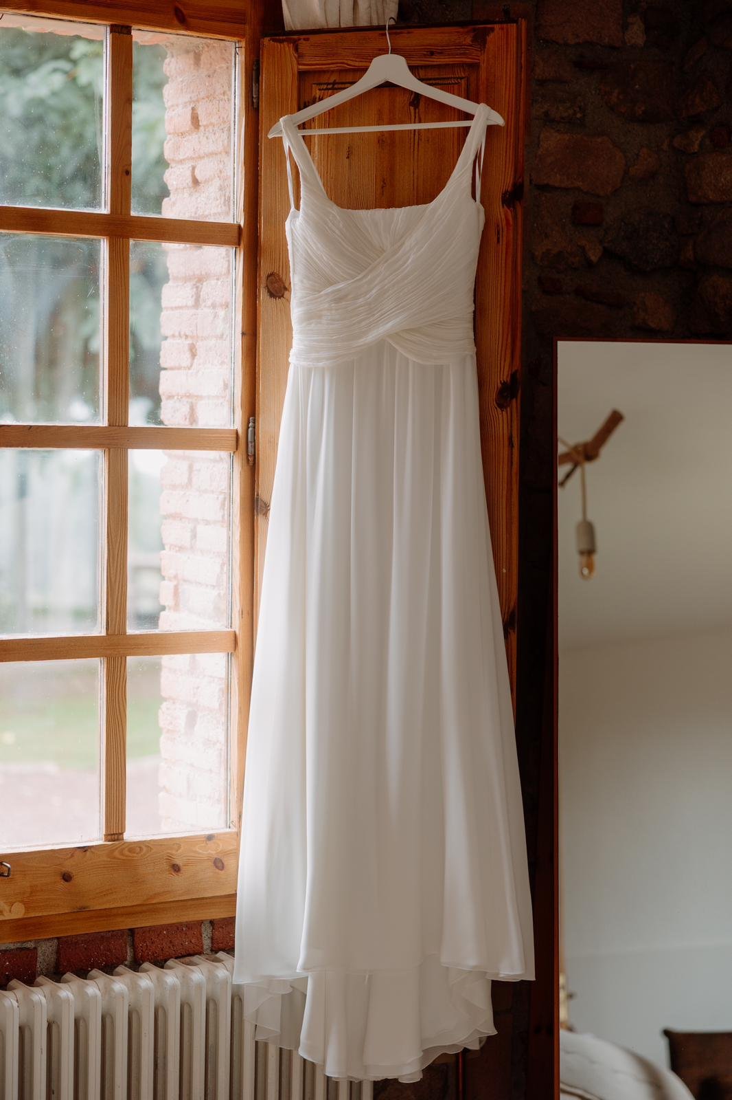 Vestido de novia de gasa de seda con cuerpo drapeado, colgado junto a una ventana
