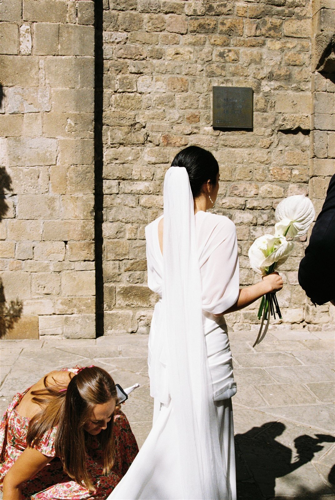 Espalda del vestido de Paula con velo largo y falda de gasa, frente a un muro de piedra en Barcelona