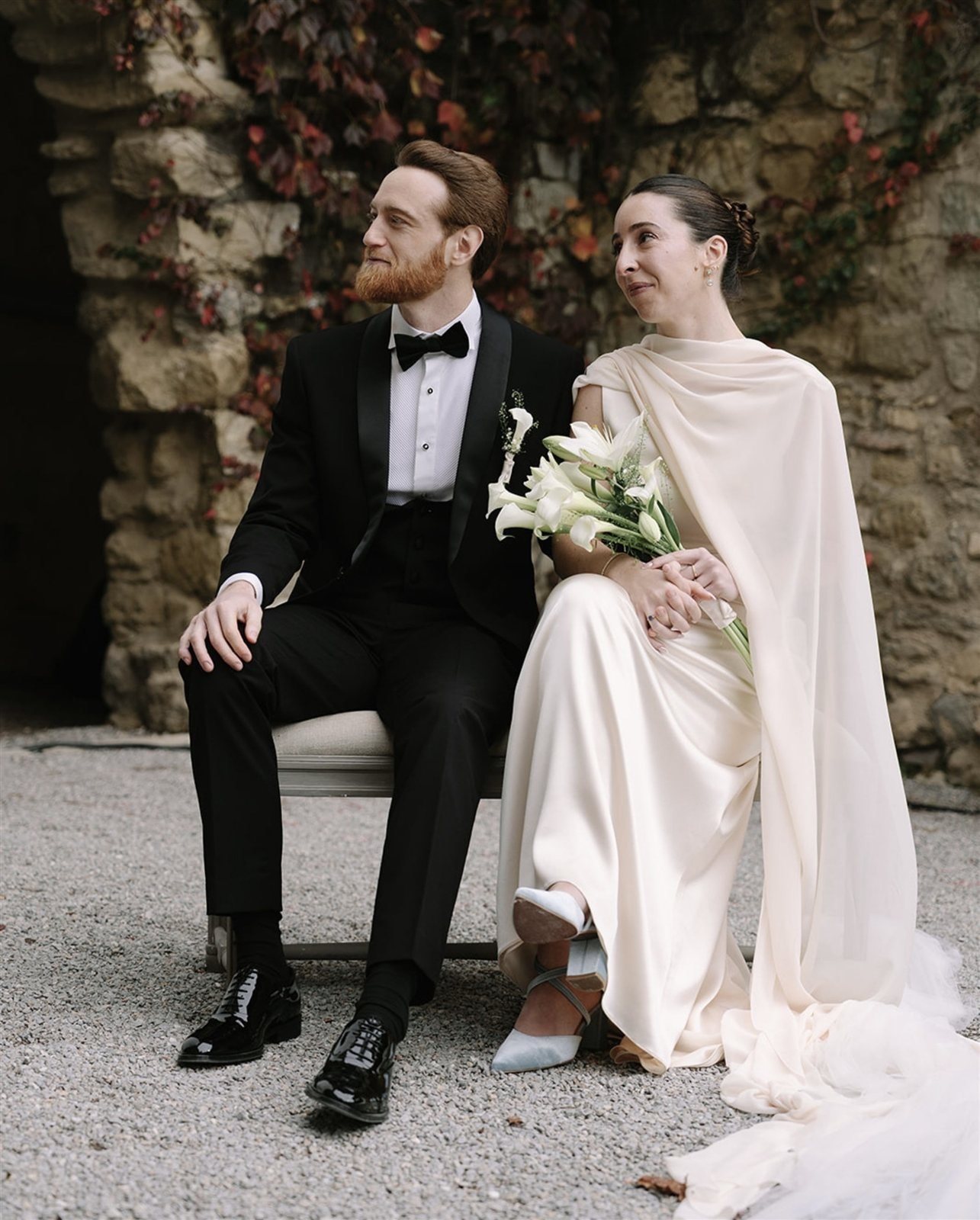 Paula y su pareja sentados, vestido de crepé satén dorado con maxichal sobre el hombro y ramo de calas